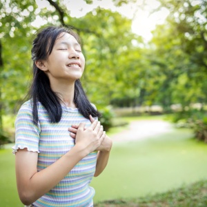 Happy smiling asian child girl standing in green nature,holding hands on heart,female teenage touching her chest,enjoy breathing fresh air with closed eyes,relaxing in park,healthy lifestyle concept