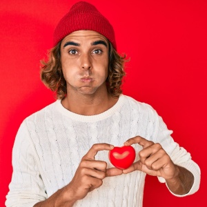 Young hispanic man holding heart puffing cheeks with funny face. mouth inflated with air, catching air.